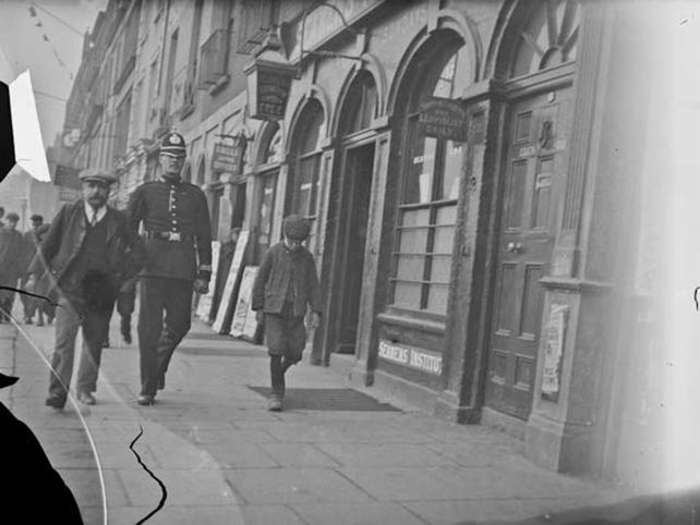 Man walking on Eden Quay accompanied by a policeman. Shops in the background include O'Dell Sons Shipping and Missions to Seamen Institute