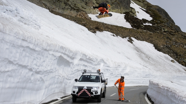 A snowboarder jumps over a road maintenance crew taking off the last snow on a mountain pass road after exceptional snowfalls in Switzerland