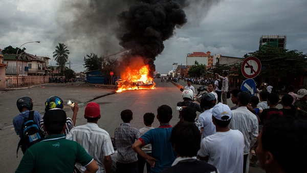 Protesters watch a burning police car in the Cambodian capital of Phnom Penh