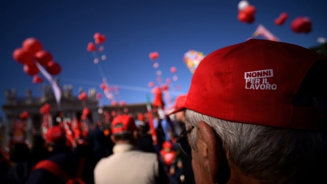 A pensioner wearing a hat reading 'Grandparents for work' stands in San Giovanni square, Rome