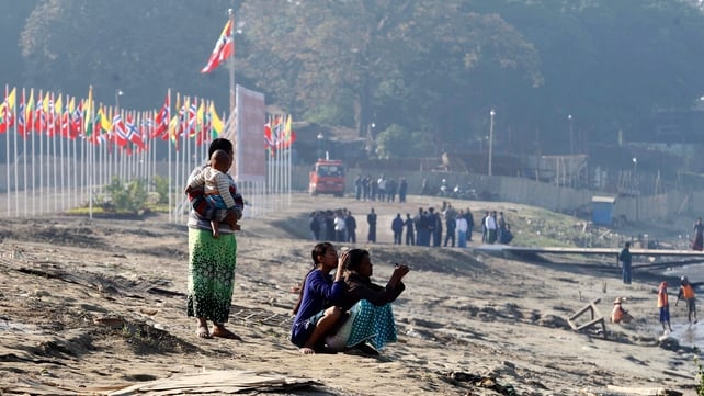 Homeless people sit on a beach as Norwegian and Myanmar flags fly to welcome Norway's king and queen near the banks of the Irrawaddy river