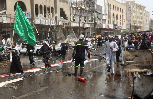 Iraqi pilgrim holding a green flag walks past destroyed cars at the scene of a car bomb blast