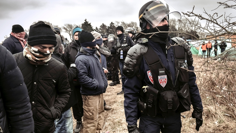Policemen stand next to agents dismantling shacks as refugees look on in the ‘Jungle’ camp