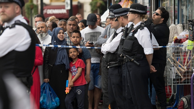 Members of the public watch police officers standing outside an address in Barking