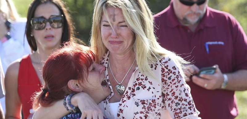 Tuismitheoirí agus daltaí ag tabhairt sóláis dá chéile taobh amuigh de Mheánscoil Marjory Stoneman Douglas.