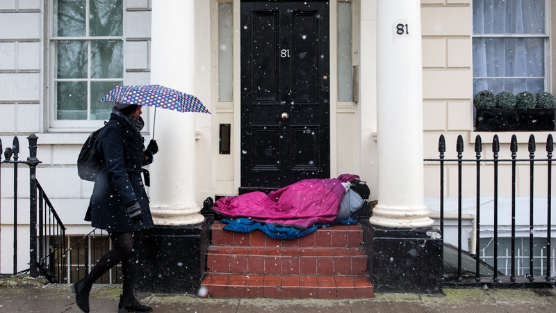 A woman walks past a homeless person sleeping in a doorway during a snow shower in London today