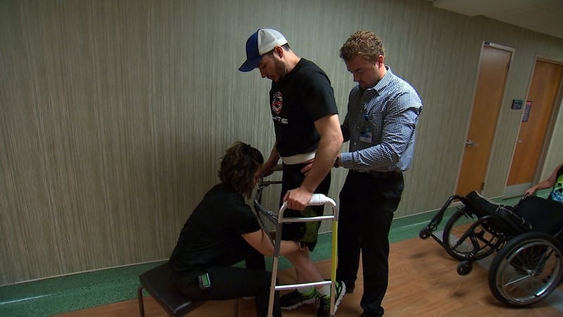 Jered Chinnock taking steps with a front-wheeled walker while trainers provide occasional assistance (Pic: Mayo Clinic)