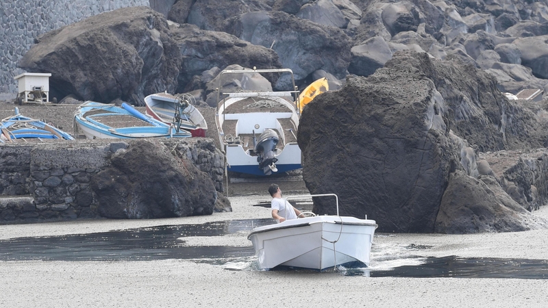 A boat leaves the port of 'Pertuso' as the coastline is seen covered with a layer of volcanic ash