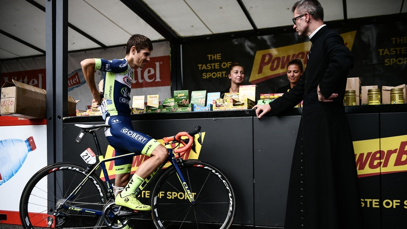 A priest looks on as France's Guillaume Martin picks up snacks at the start village in Lourdes during the 2018 Tour de France. Photo: Philippe Lopez/AFP via Getty Images