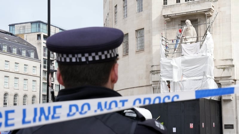 A police officer watches as a man stands of scaffolding surrounding the statue