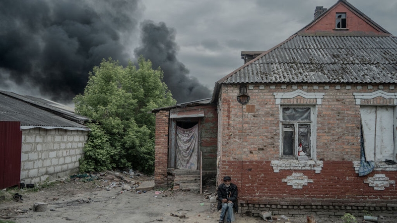 An elderly man sits outside his shell-damaged house as smoke rises over the city following Russian bombs in Kharkiv