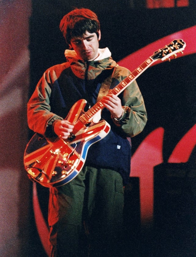 Noel Gallagher performs onstage with his Union Jack Epiphone guitar, at Maine Road Stadium in Manchester, 1996. Photo by Pete Still/Redferns.