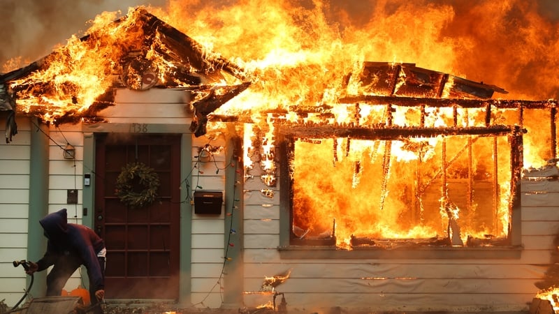 A person uses a garden hose in an effort to save a neighbour's home