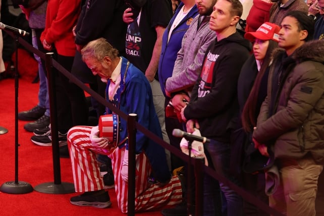 A man dressed in a costume reminiscent of Abraham Lincoln kneels as Donald Trump is inaugurated. He is flanked by other people who gathered in the Capital One Arena to watch the ceremony