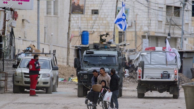 People walk along a street near Israeli military vehicles in Jenin, West Bank