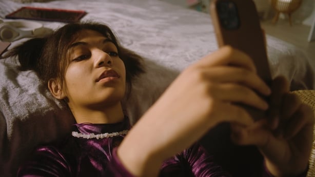 African American girl sits near bed in her room, smiles and surfs internet using phone. Beautiful teenager chats with friends or watches video content. Comfortable home with cozy and stylish interior.