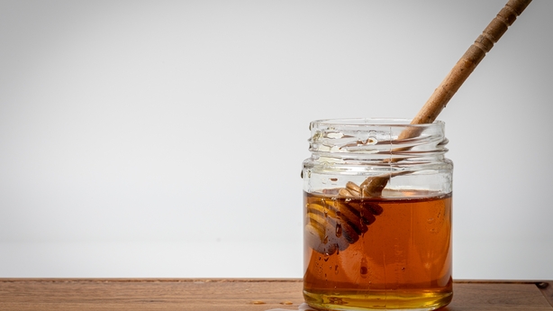 Close up photo of honey dipper immersed in a jar of honey with a white background and wooden board