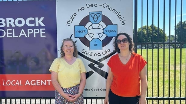 Two women stand in front of a Gaelic Games club symbol