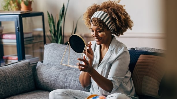 A wide angle view of a smiling African-American female holding a mirror while applying some creme on her face to keep it fresh and hydrated. She is sitting on the sofa in the living room. (skincare concept)
