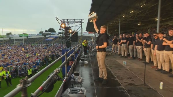A person holding the Liam McCarthy Cup on a stage at Semple Stadium with a crowd watching