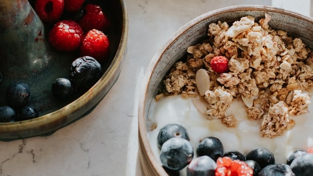A fresh and healthy breakfast spread including granola, grapes, strawberries, blueberries, raspberries and yoghurt. They are all decanted in beautiful crockery on a clean, white, marble surface.