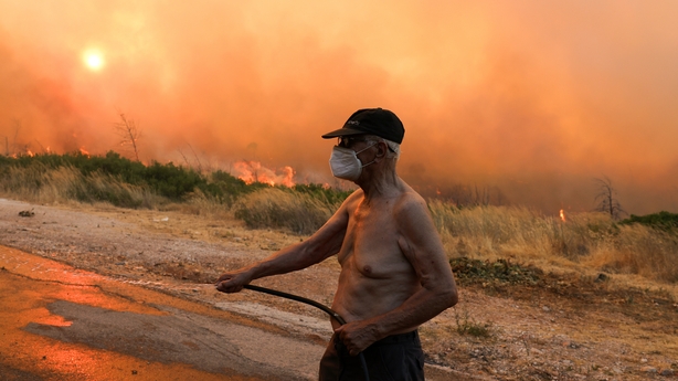 A man with no top on and wearing a mask uses a hose to put out a fire in greece with smoke billowing in the background