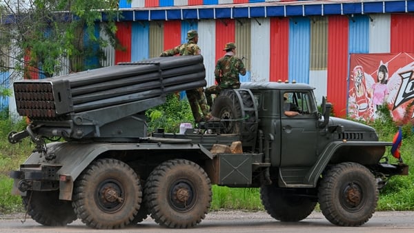 A green military vehicle with two men standing on top of it drives on a road with grassland behind it