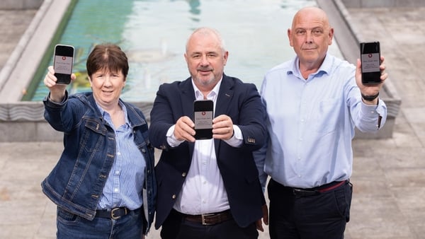 three people posing for a picture with mobile phones in their hands and a fountain in the background