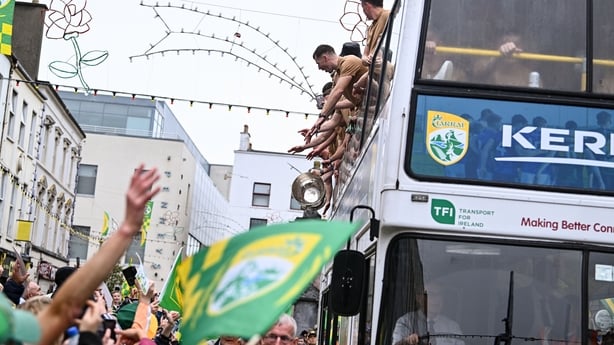 A group of people on a bus wave to people waving green and gold flags below