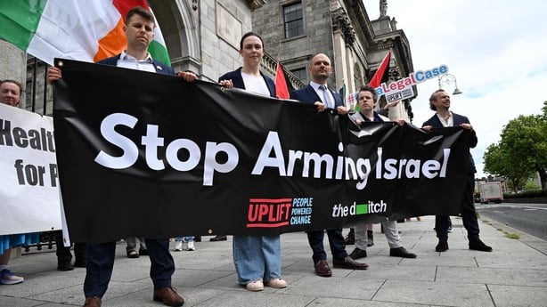 A group of men holding several banners and flags that read 'stop arming Israel' in front of a grey arch