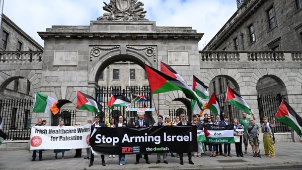 A group of men holding several banners and flags that read 'stop arming Israel' in front of a grey arch