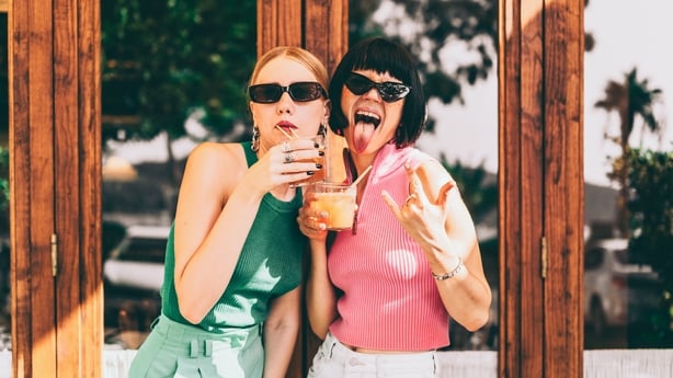 Two friends wearing fancy jewelry and sunglasses are having fun outside the cafe. Besties are drinking summer cocktails and making funny faces to the camera. Sunny vacations, bonding, and togetherness