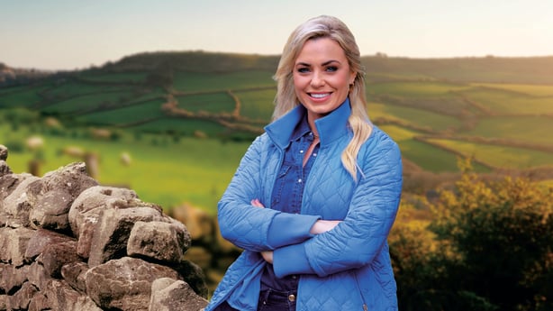 Anna Geary leaning against a stone wall smiling with her arms crossed