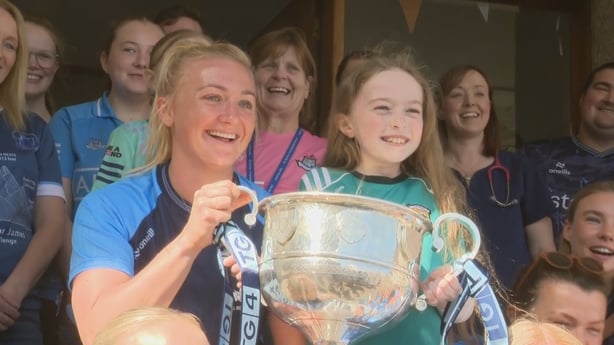 A woman with blonde hair wearing a blue jersey lifts a trophy with a young child wearing a green jersey