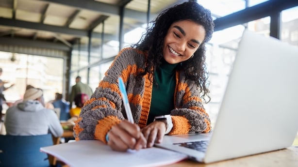 Low angle view of a smiling young female college student writing notes and working on a laptop while doing her homework in a busy campus cafeteria