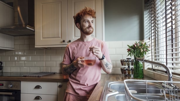 Three quarter length of a young redhead man standing in his kitchen on a morning, he is holding a cup of herbal tea and looking out the window at his home in County Durham, England. He's wearing a casual top and shorts.Video also available for this scenario.