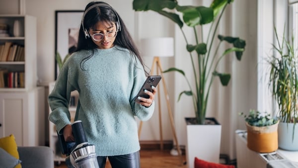 A young woman holds a cordless vacuum cleaner in one hand and a smartphone in the other in a cozy, plant-filled living room