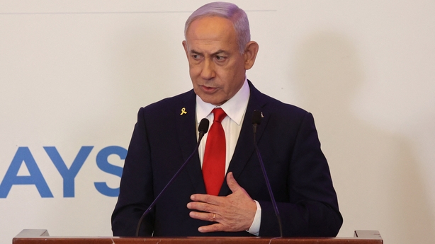 Benjamin Netanyahu wears a black suit, white shirt and red tie. He is standing facing the camera against a plain white background.