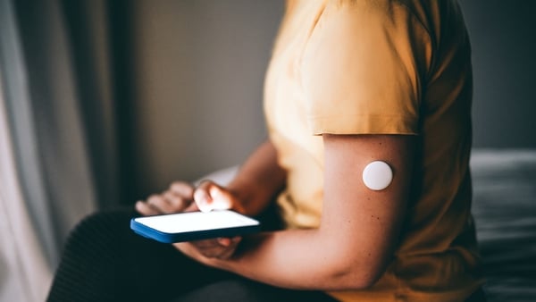 An unrecognizable person using the glucose monitor on the arm, checking the mobile app for more blood sugar level data. The wearable health sensor interacts with a smartphone application, highlighting modern health technology and remote monitoring applica
