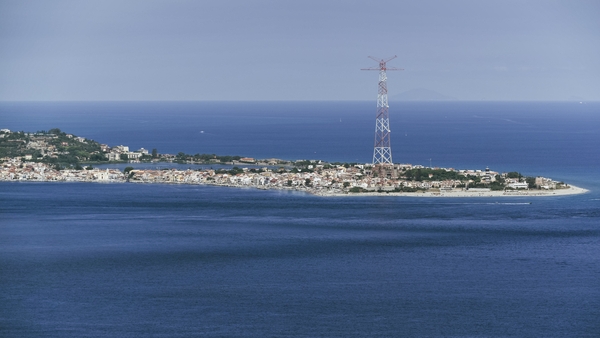 An electrical pylon is seen on a headland surrounded by the sea