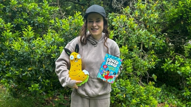 Author Aoife Dooley poses for a photo in green surroundings holding a copy of her book and toy.