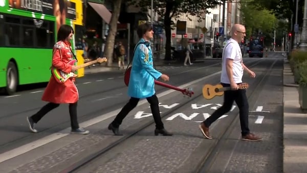 Three men, two of whom are dressed in costumes associated with ‘The Beatles’, cross a road in Dublin City centre carrying guitars
