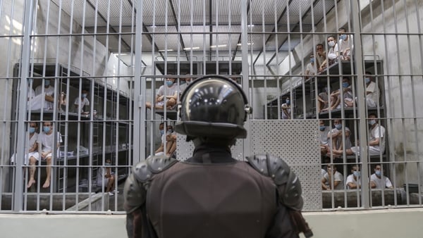 A prison officer wearing a riot helmet stands in front of a cell full of prisoners in el salvador