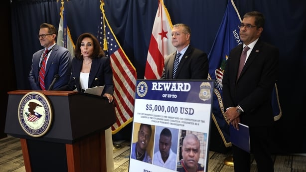 Three men and a woman stand by a lectern with an FBI wanted poster in the foreground