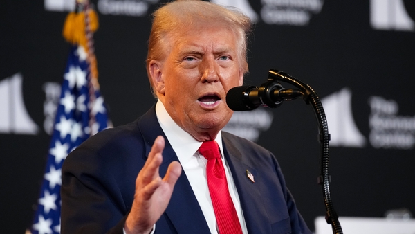 A man in a suit and red tie speaks with the american flag behind him