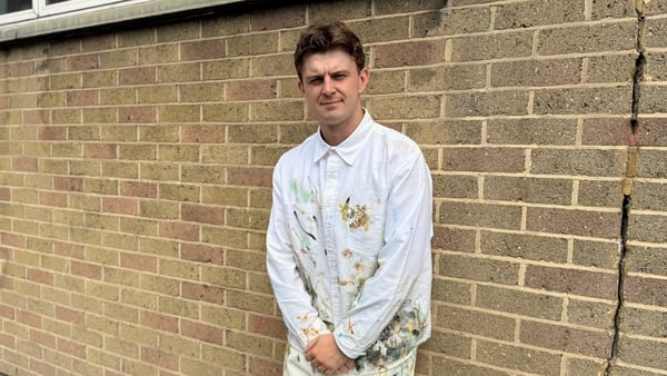 image shows Dublin artist Carl Hickey wearing white overalls covered in paint, standing against a brick wall