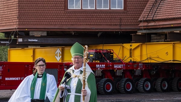 Two people dressed in clerical clothes stand in front of a building on trailers