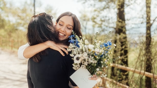 Bride holds floral bouquet, basket and envelope while hugging friend