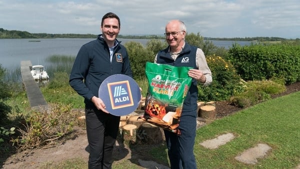 Two men standing in front of a lake with an Aldi sign and a bag of fuel