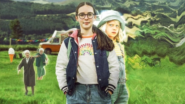 A girl stands with another girl behind her along with old ladies in the background in a green field.